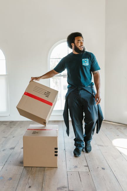 A man with dark curly hair, a beard, and medium skin tone is participating in a home relocation process within a bright, minimally decorated room. He is wearing a blue T-shirt with a logo on the chest, dark trousers, and has a jacket tied around his waist. He is holding a cardboard box wrapped with red packing tape in his right hand, preparing to place it onto another similar box on the wooden floor. Behind him, there are large, arched windows allowing natural light to illuminate the space, which features white walls and a light wooden floor. The scene depicts the packing and loading phase of a furniture transport or house removals service, with other boxes and packing materials possibly out of view. This visual represents a professional moving or removals operation, such as those provided by Removal Company Clapham, during a residential move emphasized in the context of house removals or furniture transport, aligning with the topic of packing and moving inside a property.
