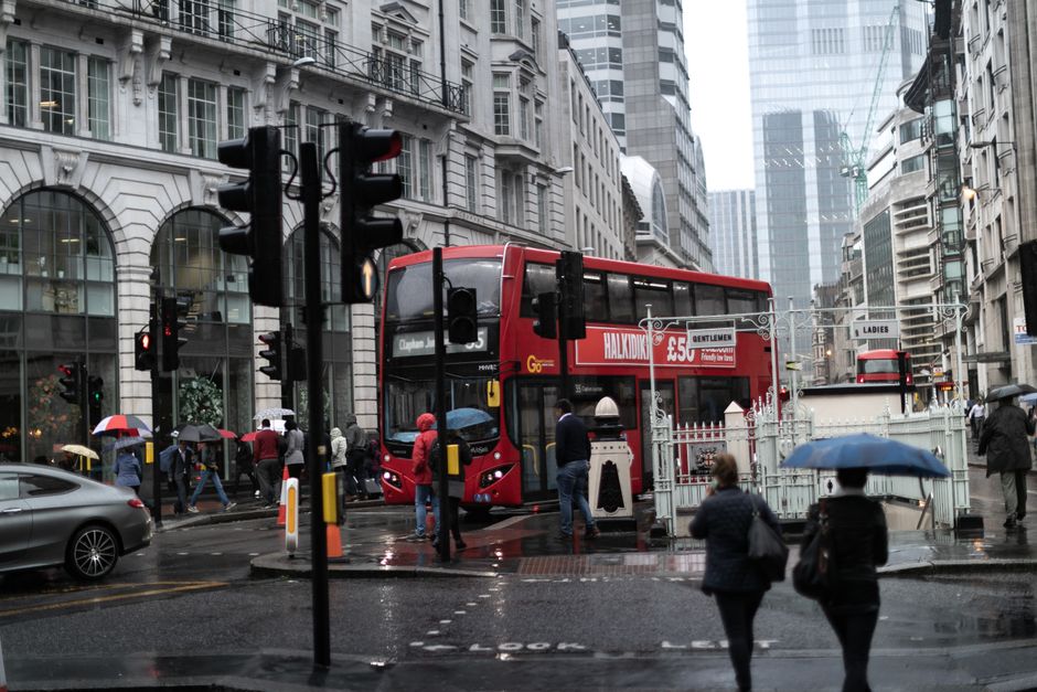 A busy street scene in central London with a red double-decker bus stopped at a pedestrian crossing, surrounded by tall modern office buildings and historic architectural structures. Several pedestrians, some holding umbrellas, walk across the wet pavement and wait near the crossing. The road features traffic lights, street signs, and a small barrier, with a nearby parked gray car and another red bus visible further along the street. The scene appears to be during rainy weather, with reflections on the wet surface and overcast skies. This image captures the typical urban environment where house removals or furniture transport may occur, illustrating a bustling city area familiar to home relocation projects in Clapham.