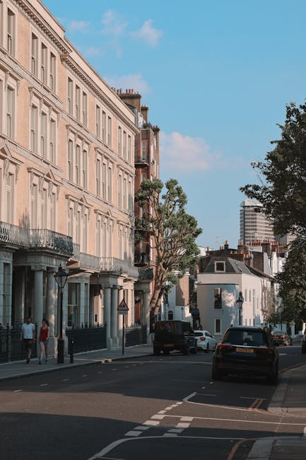 A street scene in Clapham with a row of multi-storey residential buildings on the left, featuring large windows, balconies with wrought iron railings, and classical architectural details. In the foreground, there are parked cars along the curb and a pavement wide enough for pedestrians, with a few individuals walking. Mature trees line the street, providing greenery against the backdrop of a clear blue sky with some scattered clouds. The presence of a van and other vehicles suggests ongoing home relocation or furniture transport activity near a property, which is associated with [COMPANY_NAME]’s moving services. The environment appears well-lit with natural daylight, illustrating a typical urban setting suitable for removals and packing processes involved in house removals around Clapham Common and SW4.