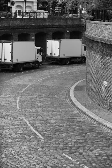 A black and white image showing two medium-sized moving trucks parked on a cobblestone street adjacent to a brick wall. The trucks are positioned parallel to each other, with the rear doors facing the viewer, indicating they are ready for a home relocation or furniture transport. The street curves gently to the right, and the trucks are located near the bend. In the background, there is an elevated parking area with a railing and some trees, suggesting an urban or residential setting. The scene appears to capture a loading process, with vehicles prepared for unloading or loading furniture, boxes, and packing materials, consistent with house removals services. The overall environment is quiet, with natural daylight illuminating the area, and the surroundings are clean and orderly, typical of professional moving logistics conducted by Removal Company Clapham.
