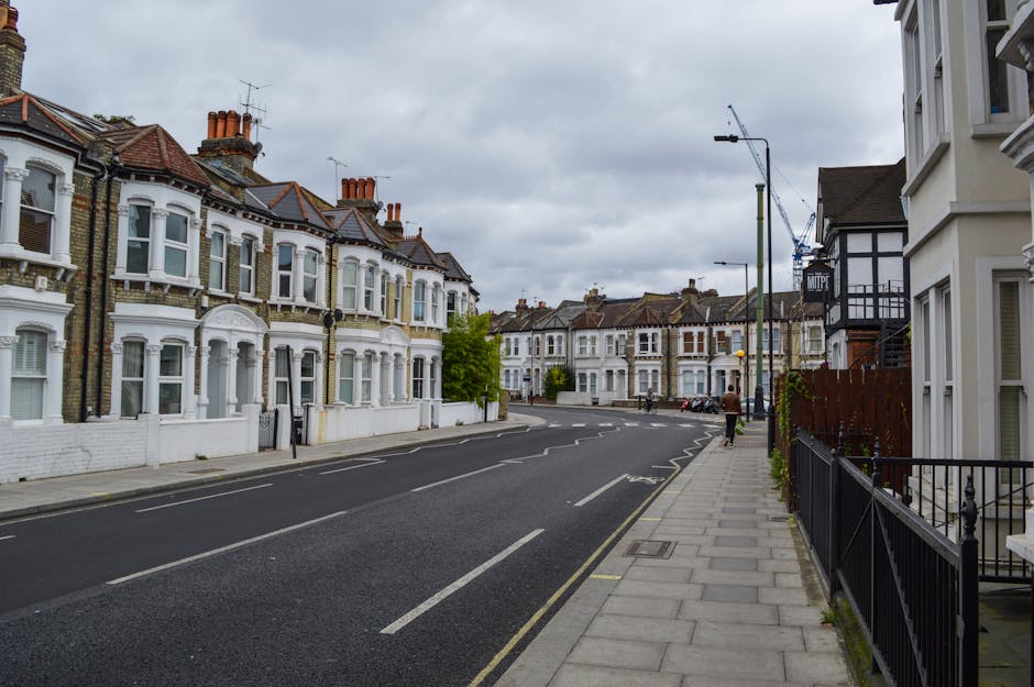 A street scene in Clapham with a row of multi-storey residential buildings on the left, featuring large windows, balconies with wrought iron railings, and classical architectural details. In the foreground, there are parked cars along the curb and a pavement wide enough for pedestrians, with a few individuals walking. Mature trees line the street, providing greenery against the backdrop of a clear blue sky with some scattered clouds. The presence of a van and other vehicles suggests ongoing home relocation or furniture transport activity near a property, which is associated with [COMPANY_NAME]’s moving services. The environment appears well-lit with natural daylight, illustrating a typical urban setting suitable for removals and packing processes involved in house removals around Clapham Common and SW4.